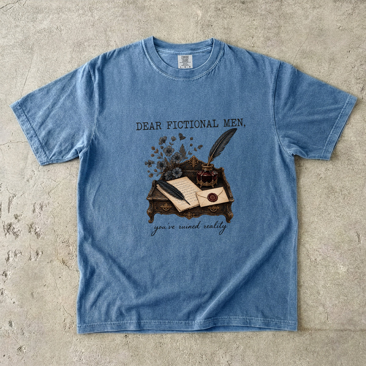 A light blue graphic t-shirt laid flat on a textured concrete surface. The shirt features a vintage-style illustration of an inkwell, quill, and open book on a desk, with the text 'DEAR FICTIONAL MEN,' above the graphic and 'you're meant really' below.