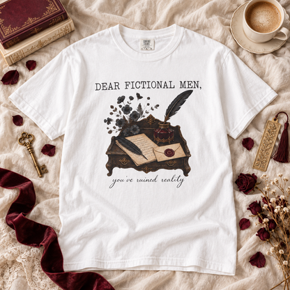 A white graphic t-shirt laid flat on a beige fabric background with vintage books, a coffee cup, dried flowers, and rose petals. The t-shirt features a dark illustration of a desk with an open book, inkwell, and feather quill, with the text 'DEAR FICTIONAL MEN, you've ruined really'.