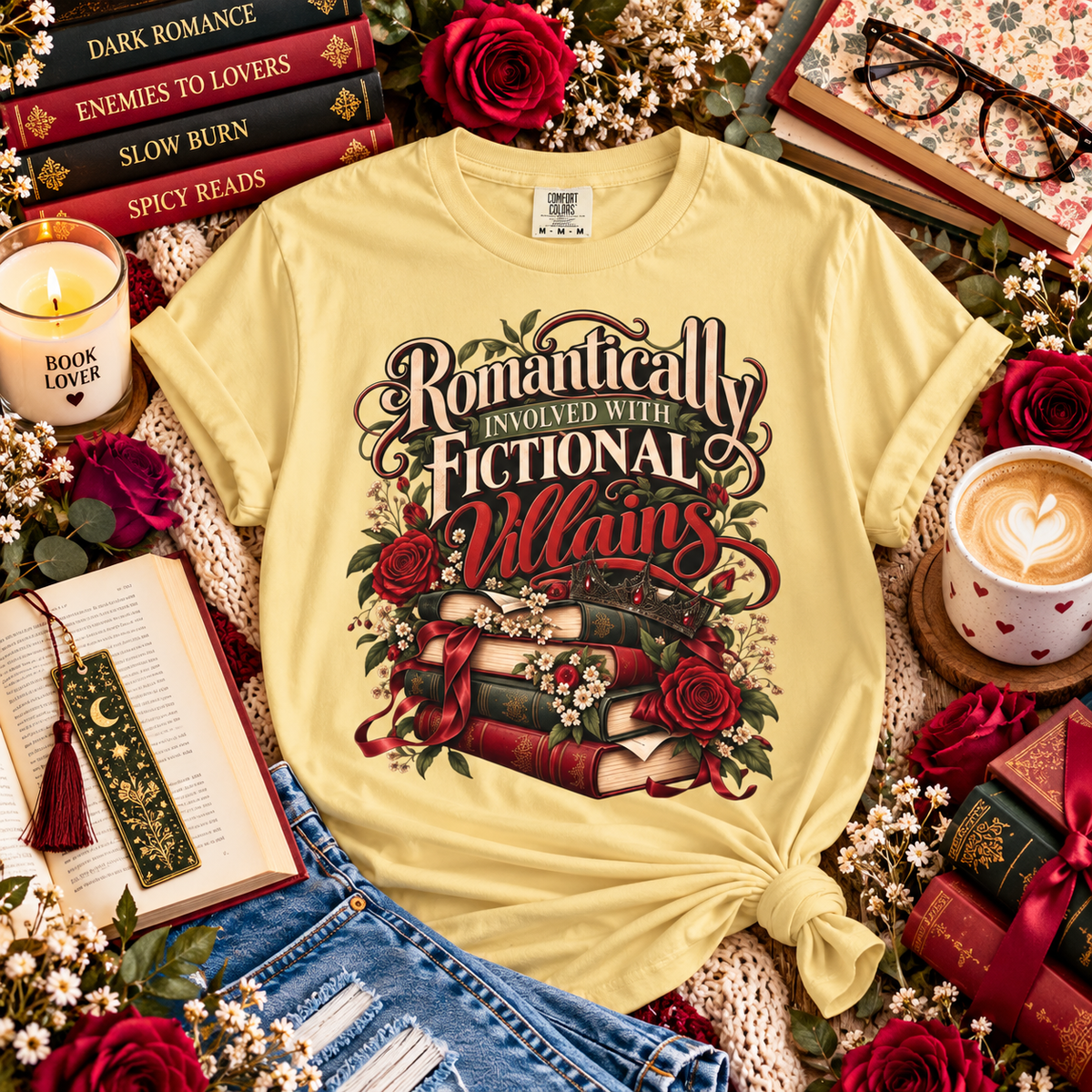 A pale yellow graphic T-shirt laid flat in a styled flat-lay arrangement with books, roses, flowers, a candle, a cup of latte, and denim shorts. The shirt has a large decorative front print reading “Romantically Involved with Fictional Villains” in dark script with red roses and stacked books.