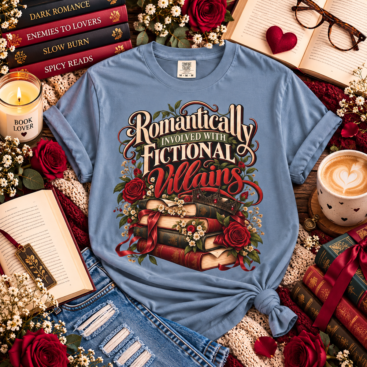 A light blue graphic T-shirt laid flat on a styled book-themed surface with roses, open books, glasses, a candle, and a cup of coffee. The shirt has a large decorative print reading “Romantically Involved with Fictional Villains” above stacked books, roses, and floral details.