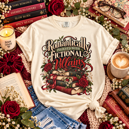 A cream-colored graphic t-shirt with rolled sleeves, styled in a cozy flat-lay arrangement with books, roses, denim, and coffee on a knitted blanket.