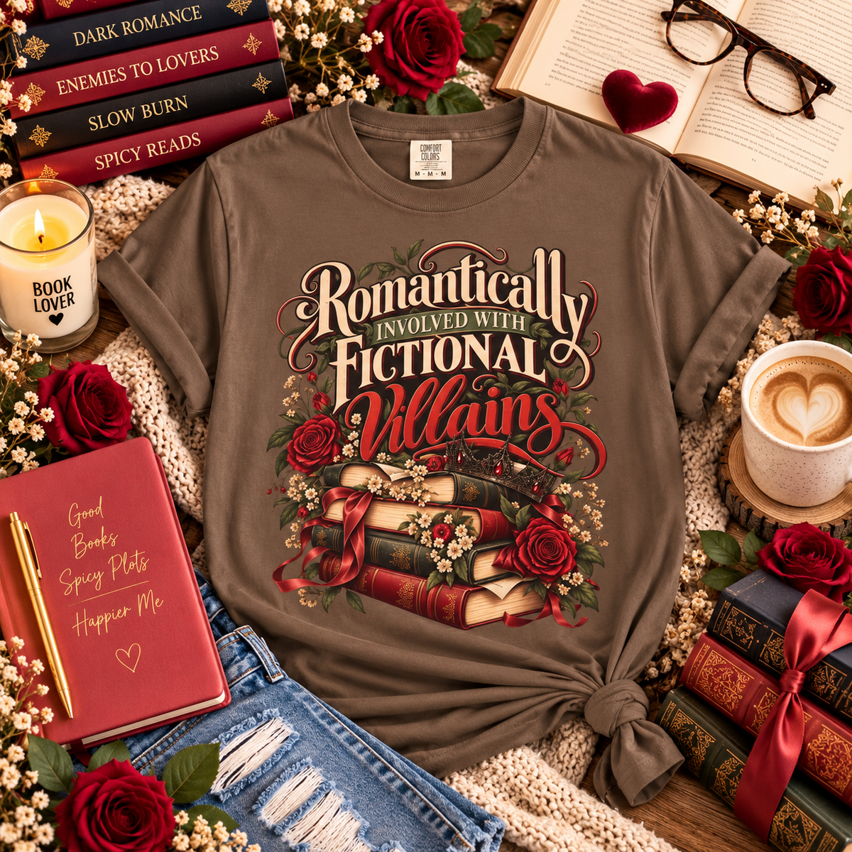 A brown graphic t-shirt with a knotted hem, featuring the text 'Romantically Involved with Fictional Villains' in a vintage font above an illustration of stacked books, roses, and a ribbon. The shirt is styled in a cozy flat-lay arrangement with red roses, a candle, a notebook, glasses, and a cup of coffee.