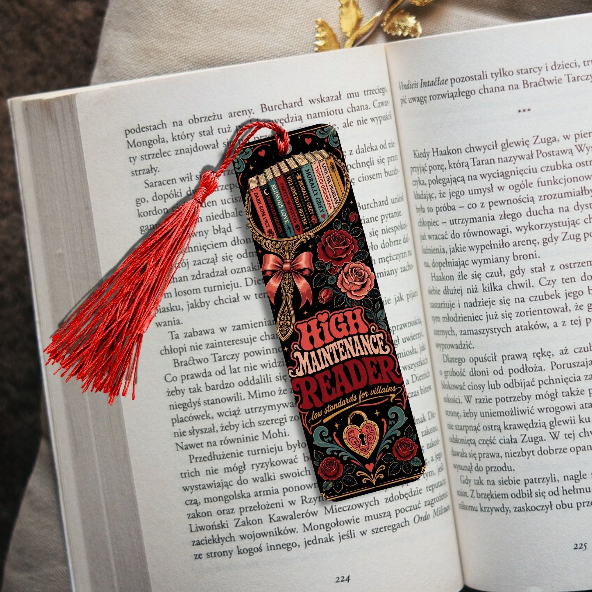 A decorative book bookmark shaped like a Bible with the title 'HIGH MAINTENANCE' and 'READER' printed on it, featuring a red tassel, resting on an open book.