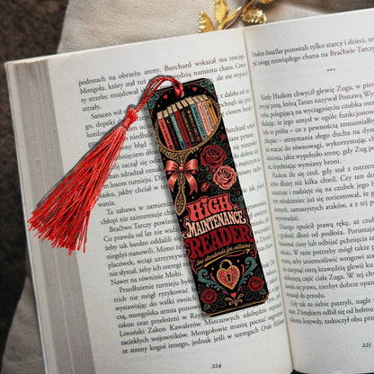 A decorative book bookmark shaped like a Bible with the title 'HIGH MAINTENANCE' and 'READER' printed on it, featuring a red tassel, resting on an open book.