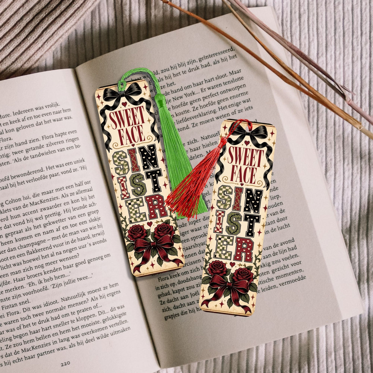 Two decorative Christmas-themed paper bookmarks or tags with 'SWEET CHRISTMAS' text, shown on top of an open book with wooden sticks. One tag has a green tassel and the other has a red tassel.