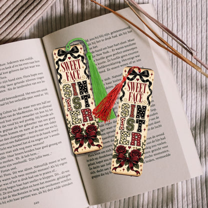 Two decorative Christmas-themed paper bookmarks or tags with 'SWEET CHRISTMAS' text, shown on top of an open book with wooden sticks. One tag has a green tassel and the other has a red tassel.
