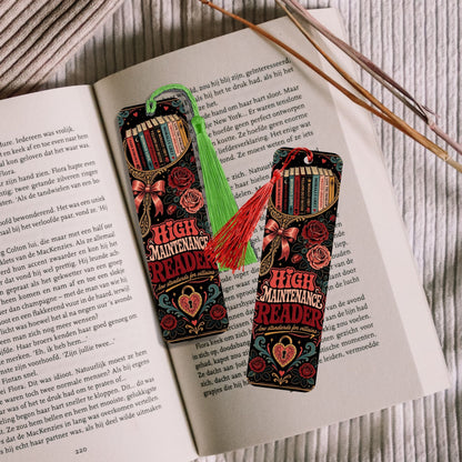 Two decorative book-shaped bookmarks with tassels resting on open book pages, with wooden sticks in the background.