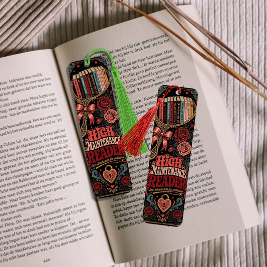 Two decorative book-shaped bookmarks with tassels resting on open book pages, with wooden sticks in the background.