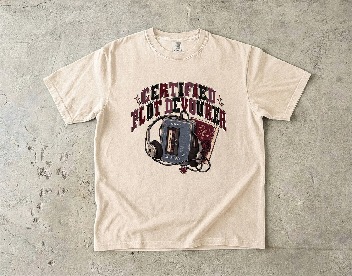 A cream-colored short-sleeve graphic t-shirt laid flat on a textured gray concrete surface. The graphic features the text 'CERTIFIED PLOT DEVOURER' in a dark red arched font above an illustration of a vintage cassette tape player, headphones, and a red book.