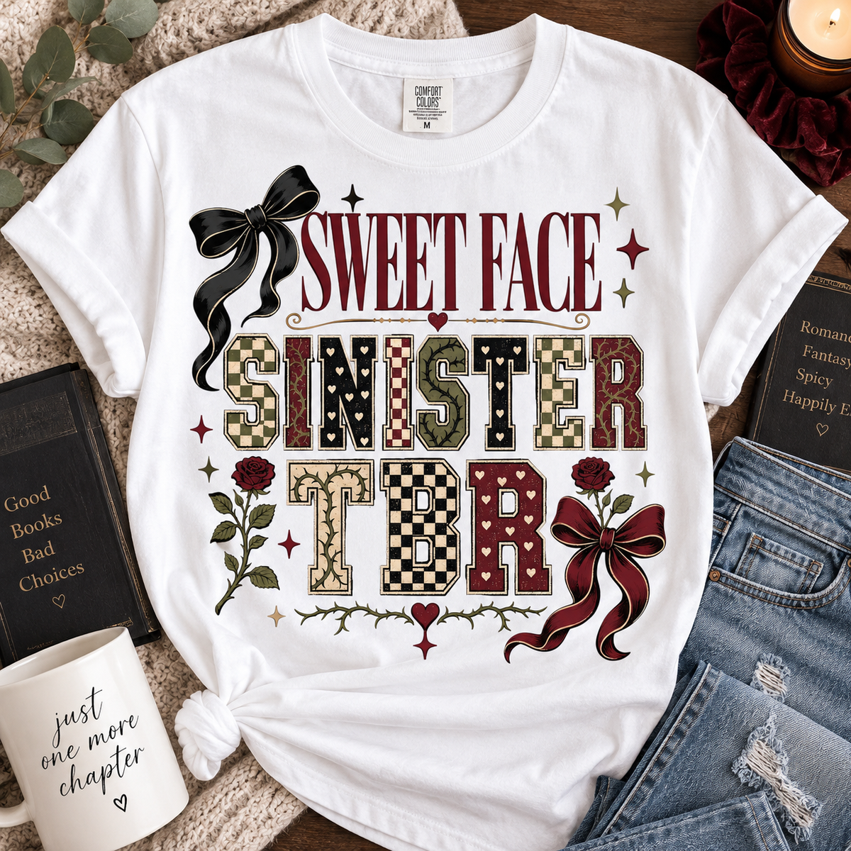 A white graphic t-shirt featuring the text 'SWEET FACE SINISTER TBR' in a vintage horror-style font, surrounded by decorative elements like a black bow, red roses, checkered and camouflage patterned letters, and small stars. The t-shirt is styled in a flat-lay arrangement with a mug, books, and denim jeans on a textured blanket background.