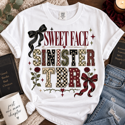A white graphic t-shirt featuring the text 'SWEET FACE SINISTER TBR' in a vintage horror-style font, surrounded by decorative elements like a black bow, red roses, checkered and camouflage patterned letters, and small stars. The t-shirt is styled in a flat-lay arrangement with a mug, books, and denim jeans on a textured blanket background.