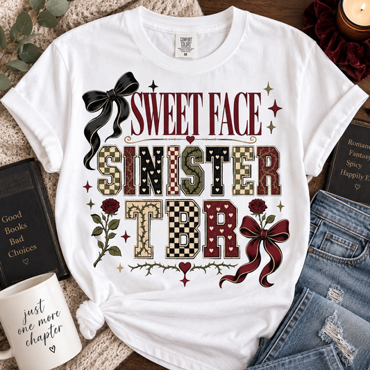 A white graphic t-shirt featuring the text 'SWEET FACE SINISTER TBR' in a vintage horror-style font, surrounded by decorative elements like a black bow, red roses, checkered and camouflage patterned letters, and small stars. The t-shirt is styled in a flat-lay arrangement with a mug, books, and denim jeans on a textured blanket background.