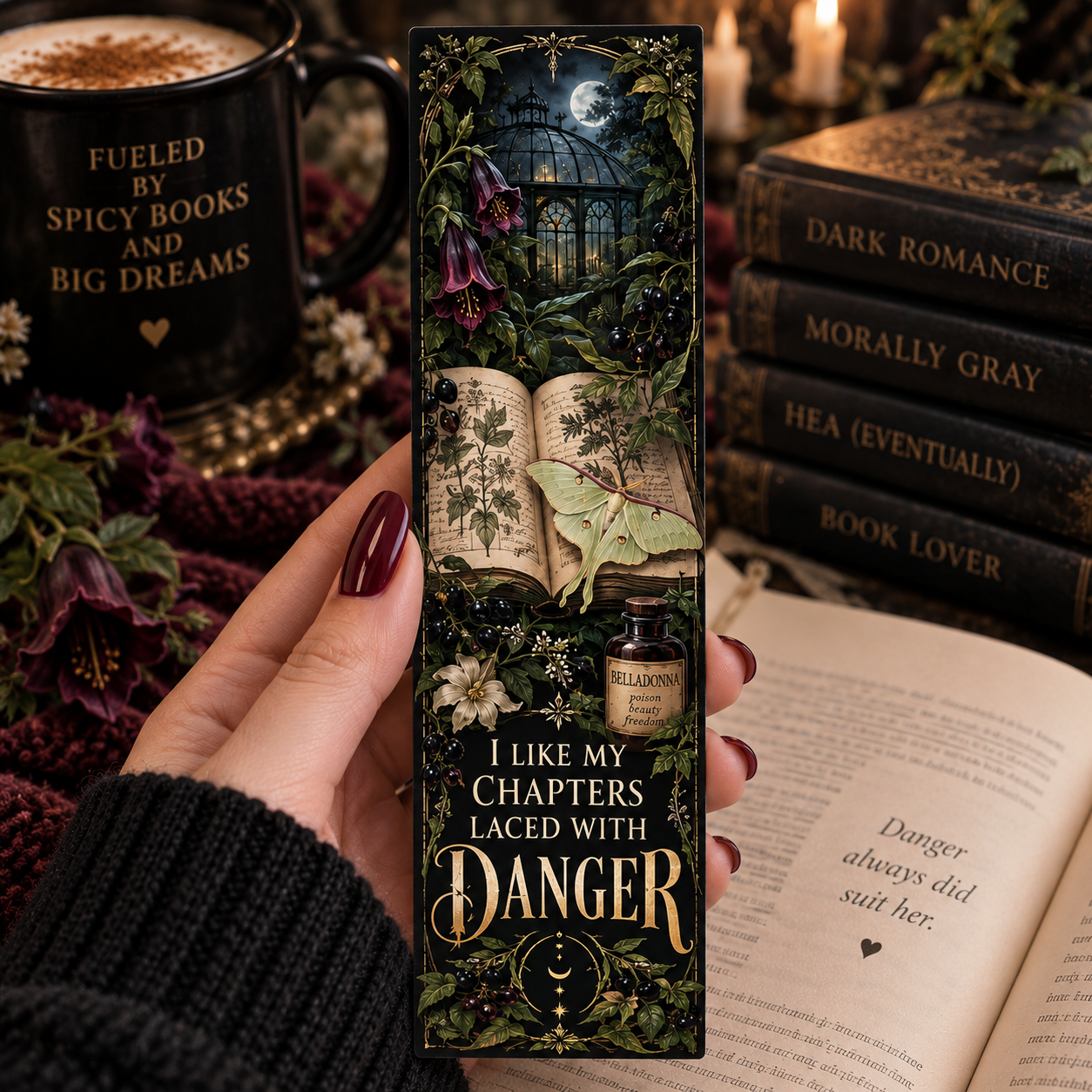 A hand holds a tall decorative book bookmark featuring a dark gothic illustration of an open book, moonlit window, flowers, and a small potion bottle. The bookmark reads, 'I LIKE MY CHAPTERS LACED WITH DANGER.