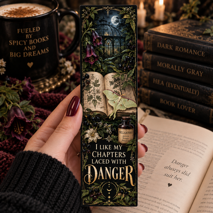 A hand holds a tall decorative book bookmark featuring a dark gothic illustration of an open book, moonlit window, flowers, and a small potion bottle. The bookmark reads, 'I LIKE MY CHAPTERS LACED WITH DANGER.